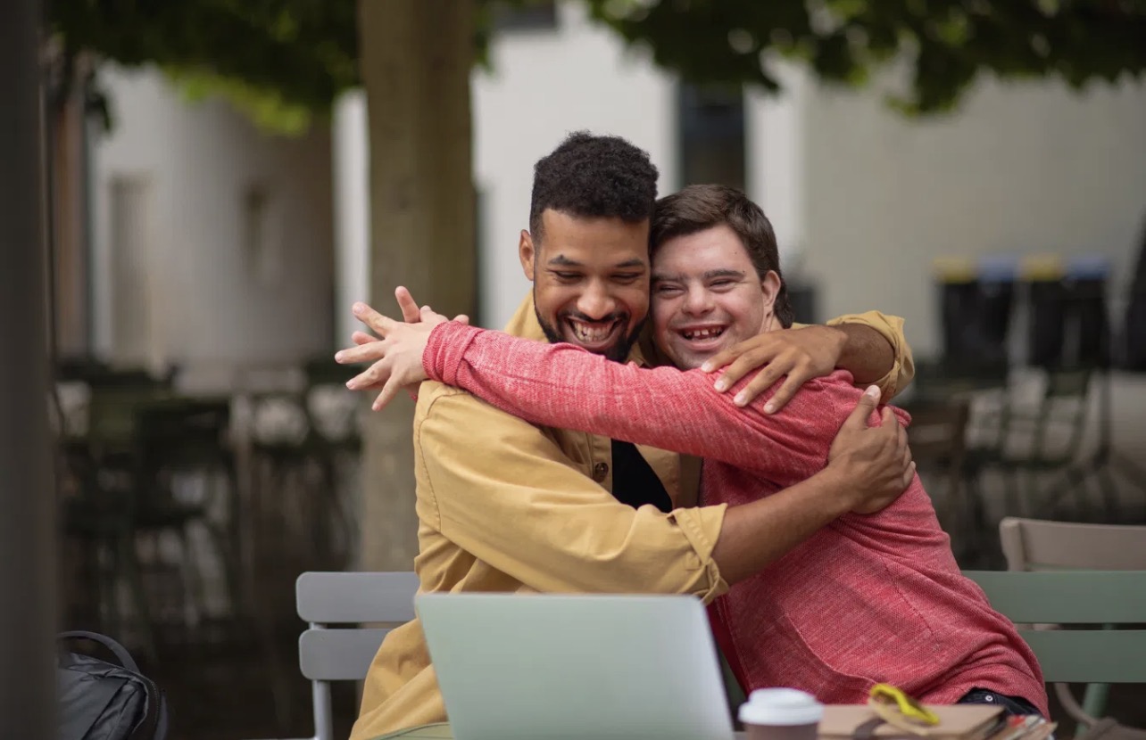 Two friends sharing a joyful hug at an outdoor cafe, one of them a young man with Down syndrome.