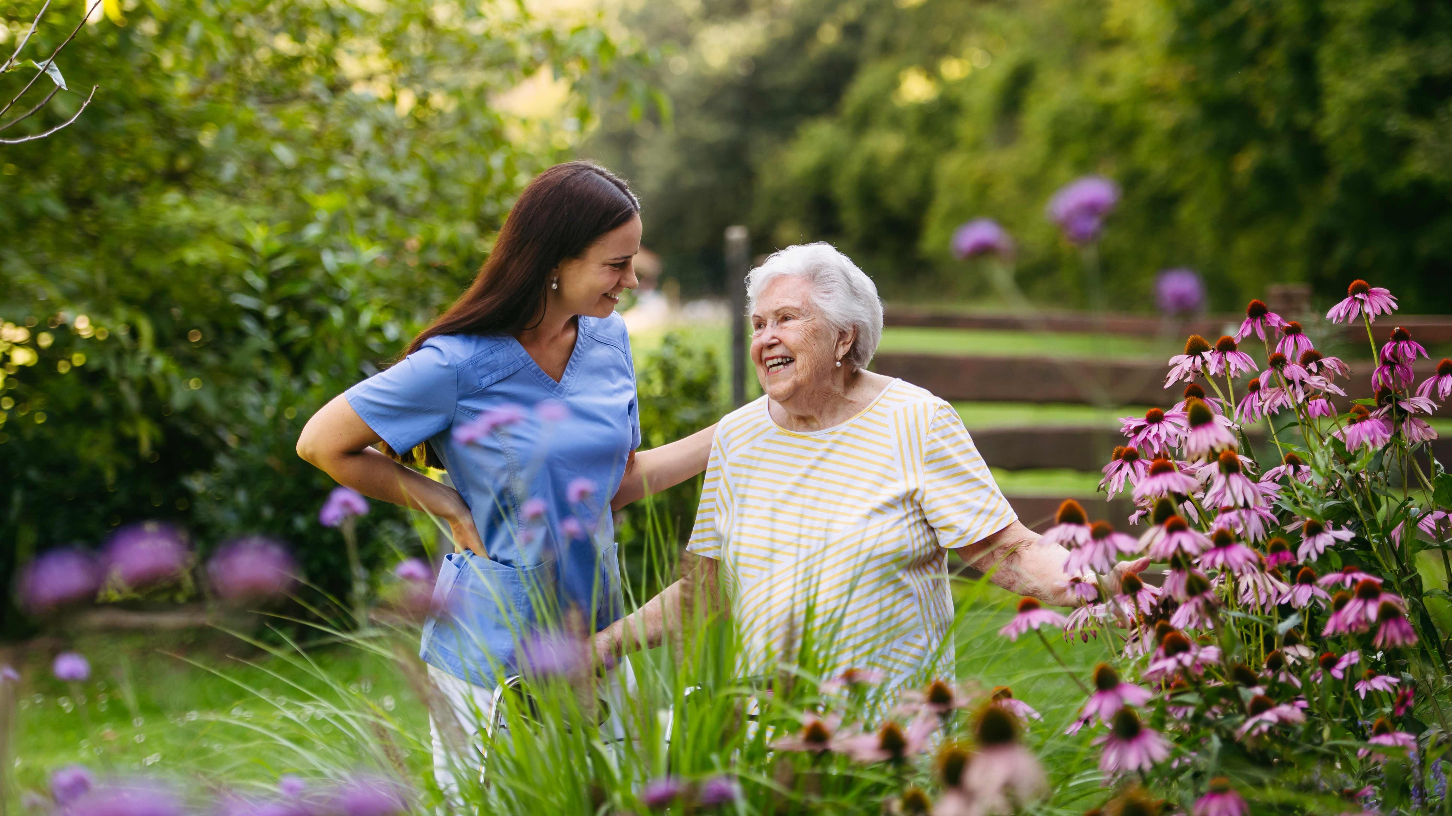 A Chriswell nurse walking beside a smiling older woman through a sunlit summer garden full of flowers.